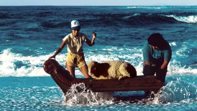 Color photo of 2 people at the shore of the Hawaiian sea, trying to sit a sheep down on a cheselong in the waves rolling onto the beach. Photo: Hipgnosis, Aubrey Powell