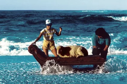 Color photo of 2 people at the shore of the Hawaiian sea, trying to sit a sheep down on a cheselong in the waves rolling onto the beach. Photo: Hipgnosis, Aubrey Powell